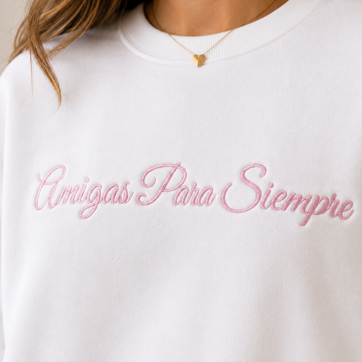 A close-up of a person wearing a white crewneck sweatshirt with 'Amigas Para Siempre' embroidered in pink script across the chest.