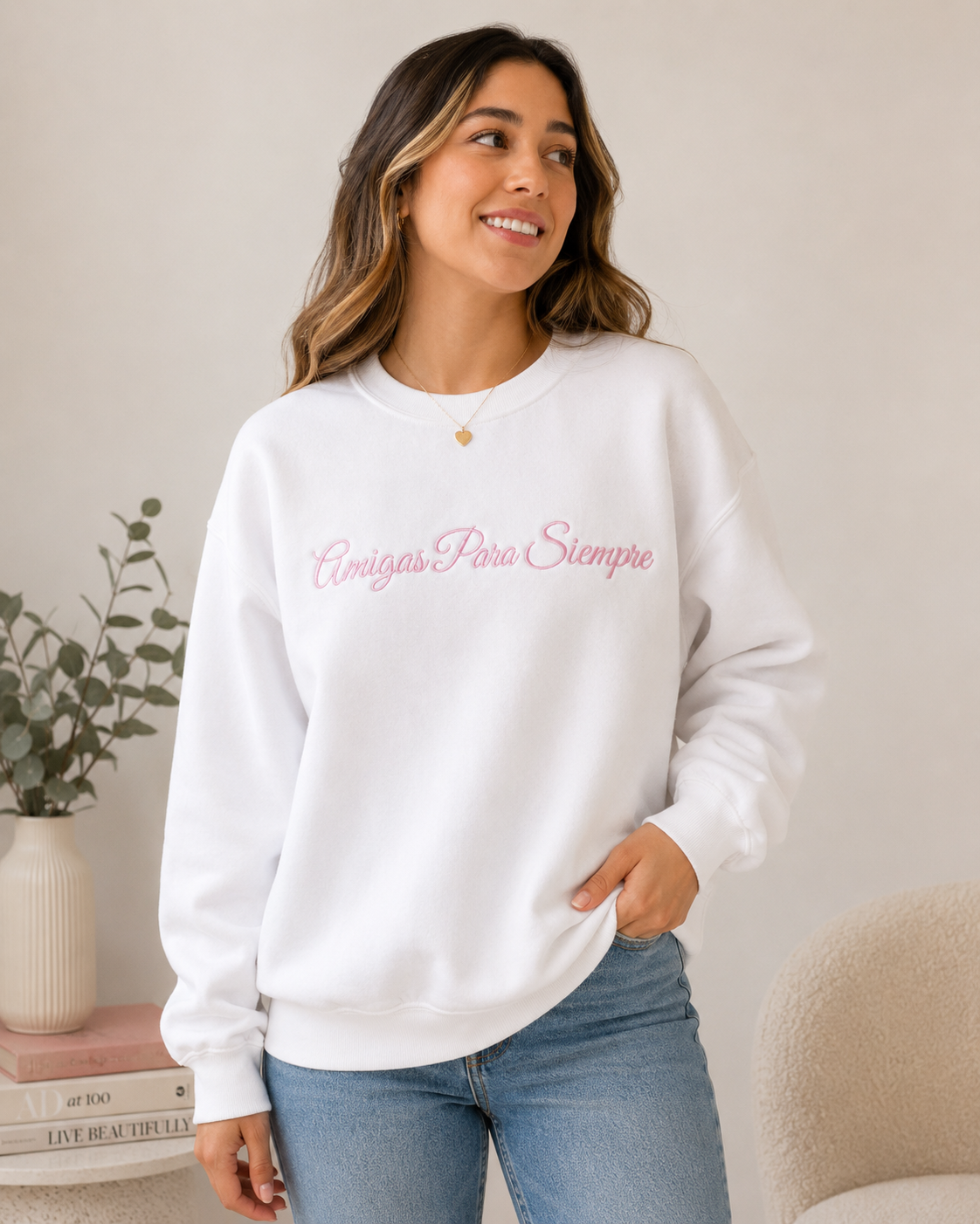 A woman wearing a white crewneck sweatshirt with pink cursive text reading 'Amigos Pero Siempre' across the chest, paired with blue jeans, standing in a brightly lit indoor setting.